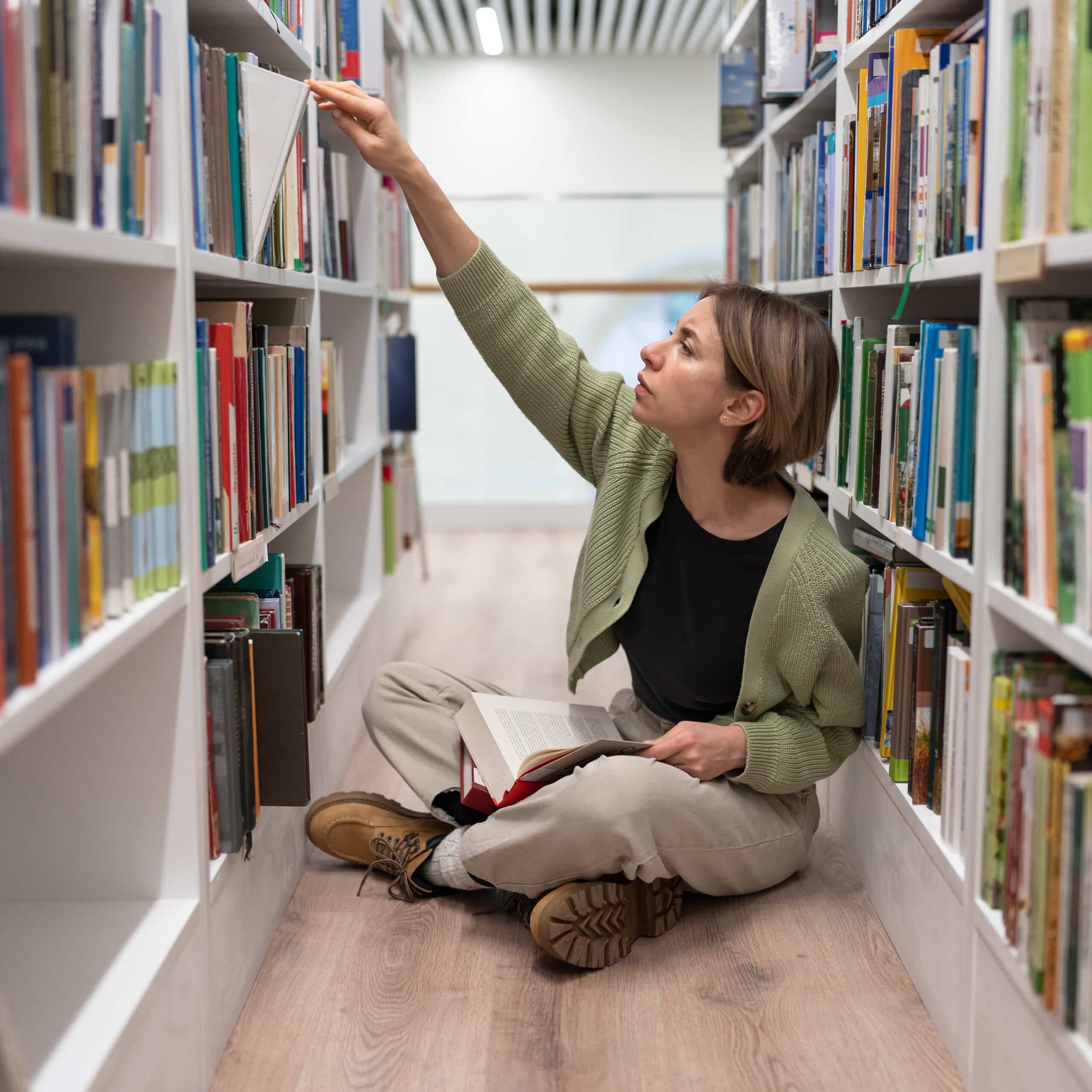 Focused female editor-in-chief of publishing house sits between bookshelves sorting books by topic.